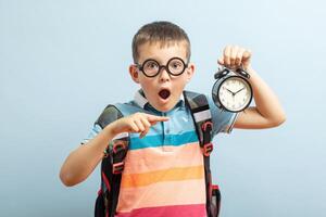 A cute school boy in glasses with an alarm clock against blue background photo
