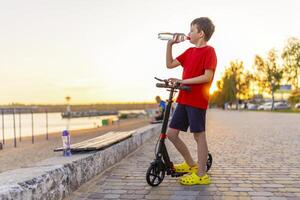 A boy drinks water from plastic bottle, standing with scooter photo