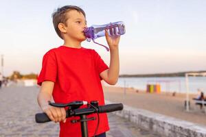 A boy drinks water from plastic bottle, standing with scooter photo