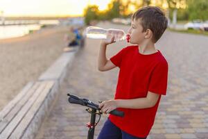 A boy drinks water from plastic bottle, standing with scooter photo