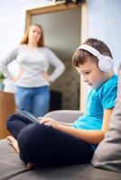 Mother scolds her son. Boy uses tablet with headphones and ignores his mom photo