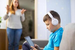 Mother scolds her son. Boy uses tablet with headphones and ignores his mom photo