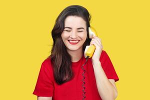 A beautiful brunette woman with red lipstick speaks on an old yellow telephone with a cord. She is smiling and happy. on a yellow background photo