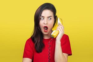 A beautiful brunette woman with red lipstick speaks on an old yellow telephone with a cord. She is shocked and surprised. photo