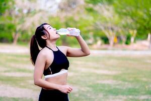 Woman hydrating post-workout in a park amidst greenery, embodying fitness and health. Teenage woman drinking water from a water bottle. Beautiful girl in black exercise clothes. photo