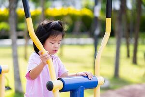 retrato de linda asiático niño aprendizaje cómo a ejercicio en corriendo ejercicio máquina a un al aire libre parque, joven niña utilizando ejercicio bicicleta en ciudad patio de juegos, en verano o primavera tiempo. niño Envejecido seis año antiguo foto