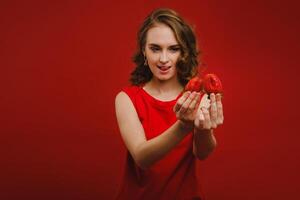 un hermosa niña en un rojo vestir en un rojo antecedentes sostiene un fresa en su manos y sonrisas foto