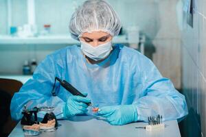 A masked and gloved dental technician works on a prosthetic tooth in his lab photo