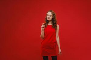 A beautiful girl in a red dress on a red background holds a strawberry in her hands and smiles photo