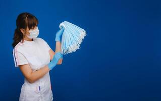 a doctor girl in a medical mask on a blue background holds a lot of protective masks in her hands photo