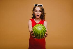 A beautiful girl in glasses and a red dress holds a watermelon in her hands photo