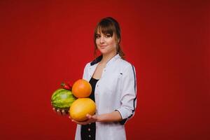 un hembra médico enfermero en un blanco Saco con Fruta en su manos poses en un rojo fondo, melón, sandía, fresa y pomelo foto