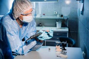 A masked and gloved dental technician works on a prosthetic tooth in his lab photo