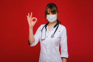 A young doctor in a white coat and medical mask stands on a red background photo