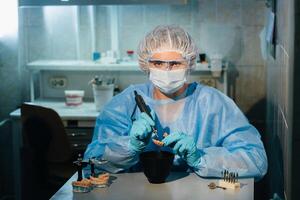 A masked and gloved dental technician works on a prosthetic tooth in his lab photo