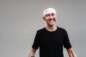 Against a gray background stands a battered and battered man in a black T-shirt with his head wrapped in a bandage photo