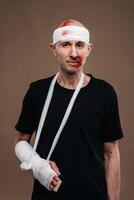 A battered man with a bandaged head and a cast on his arm stands on crutches on a gray background photo