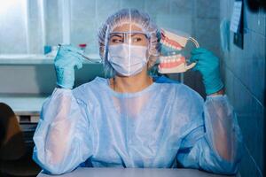 A dental doctor wearing blue gloves and a mask holds a dental model of the upper and lower jaws and a dental mirror photo