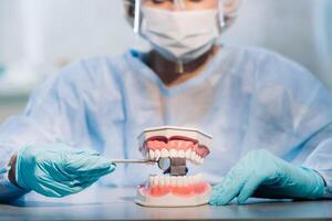 A dental doctor wearing blue gloves and a mask holds a dental model of the upper and lower jaws and a dental mirror photo