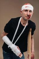 A battered man with a bandaged head and a cast on his arm stands on crutches on a gray background photo