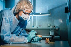 A masked and gloved dental technician works on a prosthetic tooth in his lab photo