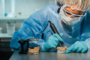 A masked and gloved dental technician works on a prosthetic tooth in his lab photo