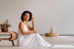 un mujer en un blanco vestido de verano en un brillante interior es sentado en un blanco alfombra. el concepto de relajación foto