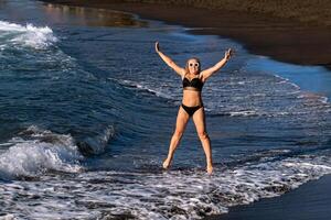 un niña en un negro traje de baño saltos en el playa en el isla de tenerife en el atlántico océano, España. foto