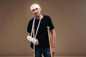 A battered man with a bandaged head and a cast on his arm stands on crutches on a gray background photo