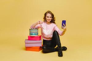 A happy young woman with colorful paper boxes after shopping, sitting and taking selfies isolated on an orange Studio background. Seasonal sales, purchases, spending money on gifts photo