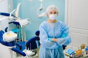 un hembra dentista vistiendo un médico máscara y caucho guantes poses para el cámara y pliegues su brazos en su oficina foto