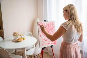 Beautiful young woman artist with palette and brush painting abstract pink picture on canvas at home. Art and creativity concept photo