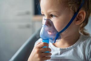 cerca arriba ver de pequeño niña son sentado y participación un nebulizador máscara propensión en contra el rostro, vías respiratorias tratamiento concepto. alto calidad foto