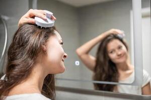 Young woman doing self hair scalp massage with scalp massager or hair brush for hair growth stimulating at home bathroom. Reflected view of the mirror. High quality photo