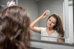 Young woman with dark hair doing self hair scalp massage with scalp massager or hair brush for hair growth stimulating at home bathroom. Reflected view of the mirror. High quality photo