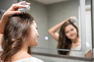 Young woman doing self hair scalp massage with scalp massager or hair brush for hair growth stimulating at home. Reflected view of the mirror photo