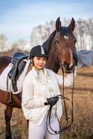 Portrait of pretty blond professional female jockey standing near brown horse in field. Friendship with horse concept photo