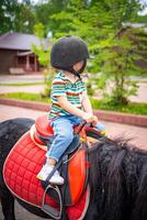 hermosa pequeño niña dos años antiguo montando poni caballo en grande la seguridad jockey casco posando al aire libre en campo foto