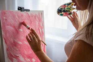 Close up of young woman artist with palette and brush painting abstract pink picture on canvas at home. Art and creativity concept photo