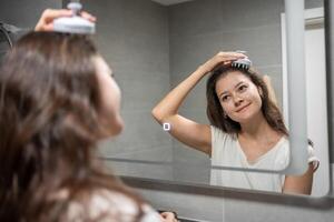 Young woman with dark curly hair doing self hair scalp massage with scalp massager or hair brush for hair growth stimulating at home bathroom. Reflected view of the mirror photo