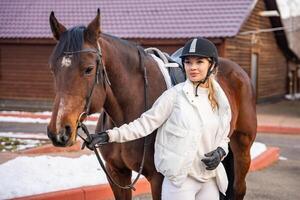 Blond professional female jockey standing near brown horse on farm in winter. Friendship with horse concept photo