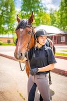 encantador joven mujer vistiendo casco acariciando a su marrón caballo foto