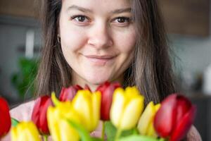 Close up portrait of smiling young woman with a bouquet of red and yellow tulips at home. Congratulations and a gift on International Women's Day or Mother's Day photo