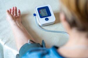 Senior woman checking blood pressure level, older female suffering from high blood pressure, sitting and using a pulsometer, tonometer. Back view photo