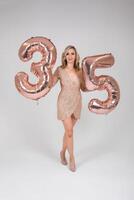 Pretty sexy blonde woman on white background. Portrait of beautiful young lady in beige glitter sequins long evening dress with thirty-five shape inflatable balloons in hand. photo