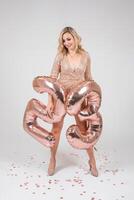 Pretty sexy blonde woman on white background. Portrait of beautiful young lady in beige glitter sequins long evening dress with twenty five shape inflatable balloons in hand. photo