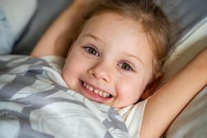 linda pequeño niña extensión su brazos felizmente con un sonrisa desde despertar arriba en su cama después siesta. alto calidad foto