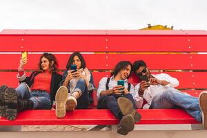 grupo de multicultural mejor amigos sentado en un grande rojo banco utilizar inteligente Los telefonos foto