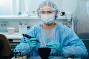 A masked and gloved dental technician works on a prosthetic tooth in his lab photo