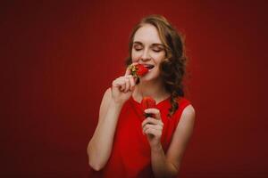 un hermosa niña en un rojo vestir en un rojo antecedentes sostiene un fresa en su manos y sonrisas foto